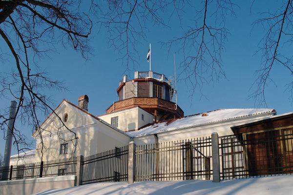Tarton tähtitorni lumisena talvena, tornissa liehumassa sini-musta-valkoinen lippu