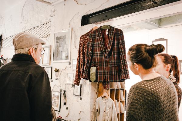 Studio shop Karud ja Pojad visitors, two women and one man exploring a vintage men's jacket and blouse. The photo also includes a wool sweater with a rabbit for a child and bear graphics on the wall.