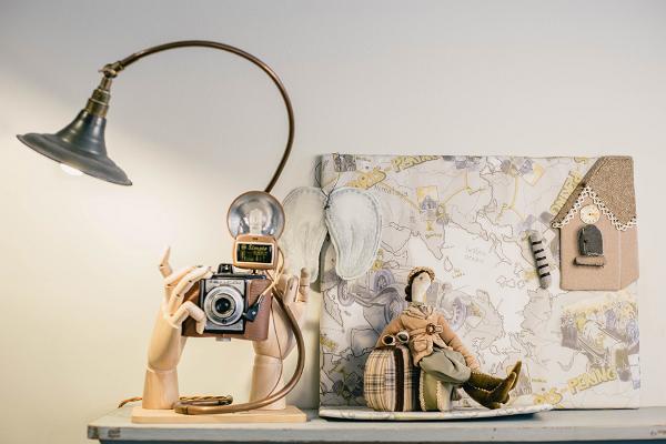 Studiobutiken Karud ja Pojad (Björnar och Söner) inifrån. Ett textilverk på hyllan (en tygdocka med väskor, en resande dam framför en karta på gränsen) och en designlampa tillverkad av en gammal fotoapparat i trähänder.