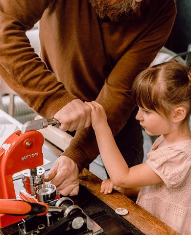 Mies ja pieni tyttö valmistavat rintamerkkiä punaisella merkkikoneella studiomyymälä Karud ja Pojadissa