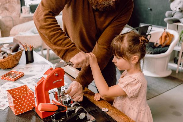 Vīrietis un maza meitene izgatavo nozīmīti studijā-veikalā "Karud ja Pojad" ar sarkanu nozīmīšu izgatavošanas mašīnu