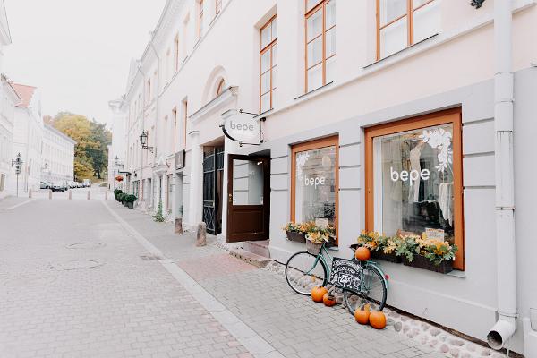 wide street view of the Bepe store, the door of the store is open, the bicycle is parked on the sidewalk next to the store and decorated with pumpkins, there are colorful autumn leaves in the flower boxes in the windows